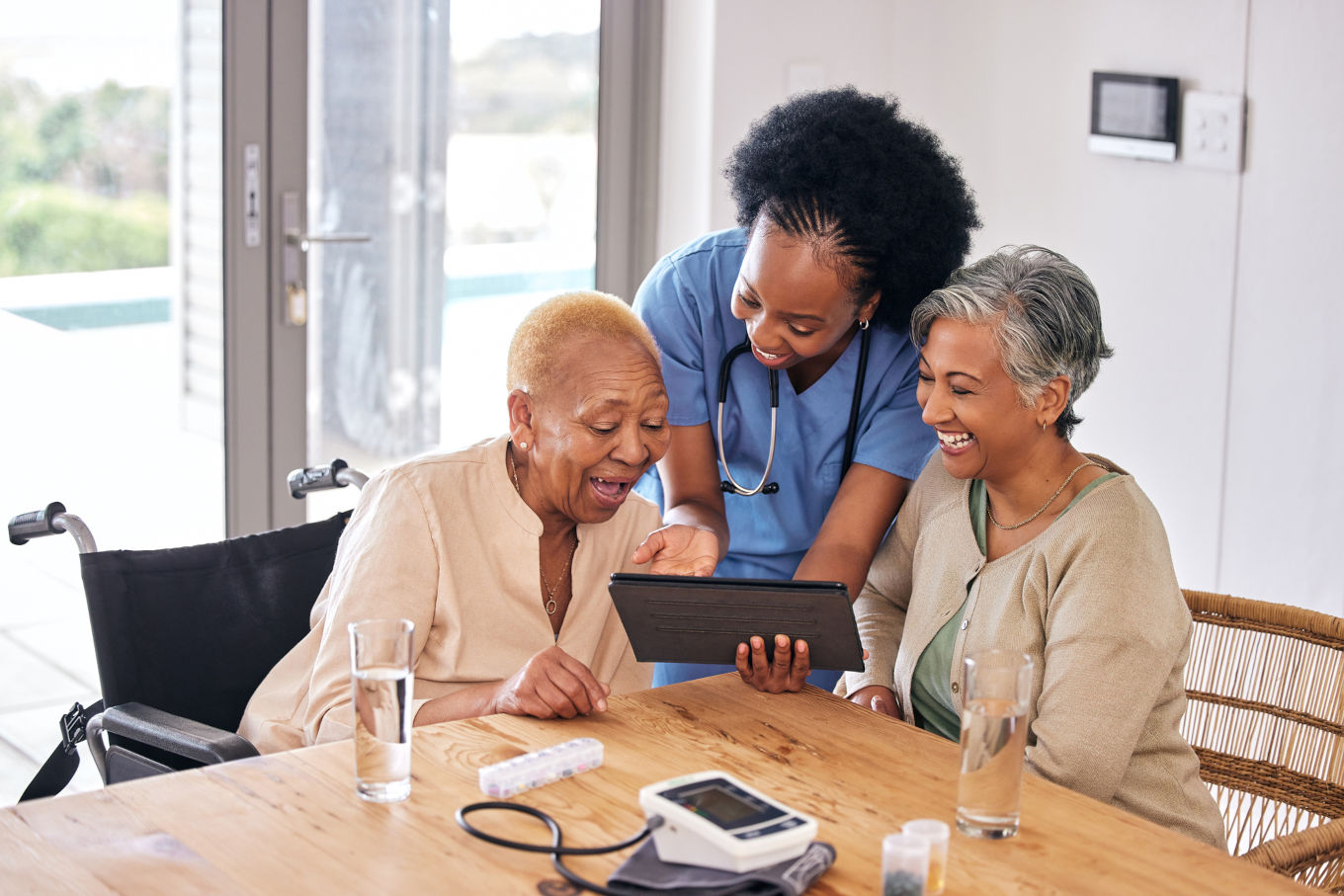 Three women, one in a wheelchair, one in scrubs, and another sitting at a table, smiling and looking at a tablet device. A blood pressure monitor and glasses of water are on the table.