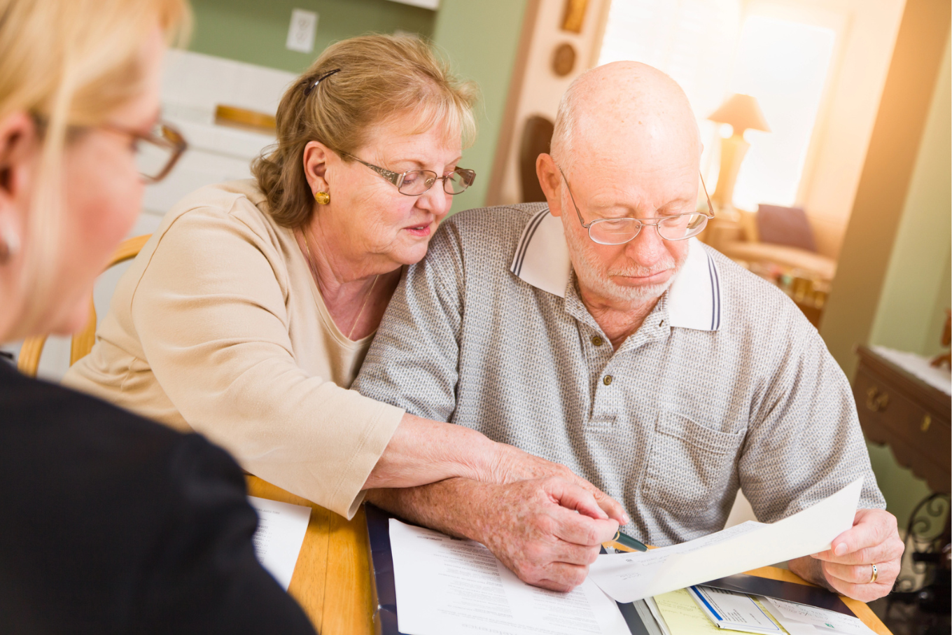 An elderly couple reviews documents at a table while another person sits nearby. The couple appears focused and engaged as they examine the paperwork.