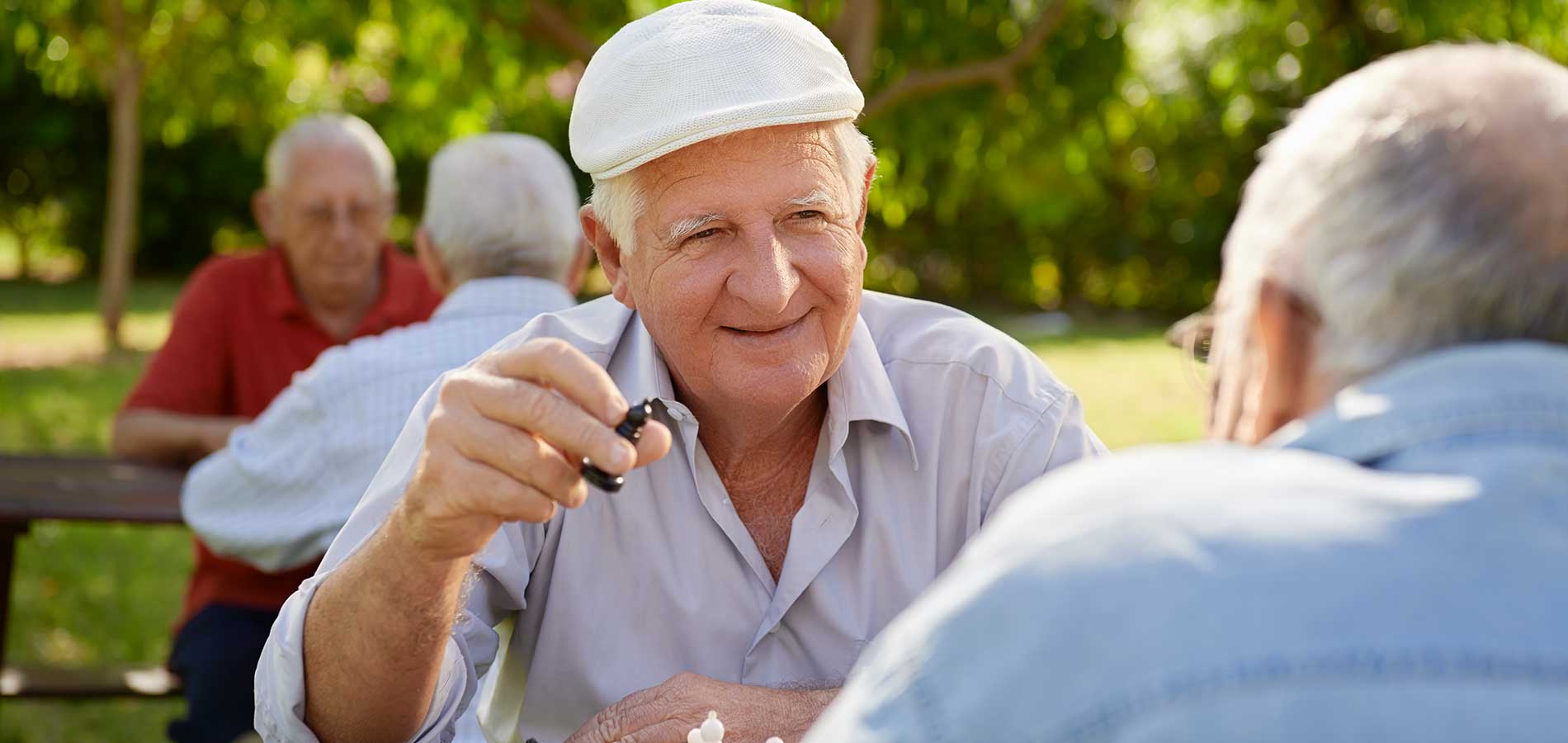 Elderly man with a flat cap playing chess in the park, smiling as he makes a move, with another man and a third person in the background.