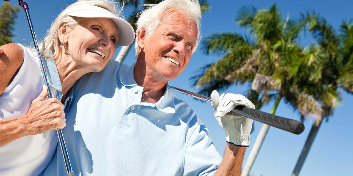 Senior couple smiling and holding a golf club together under a sunny sky with palm trees in the background.