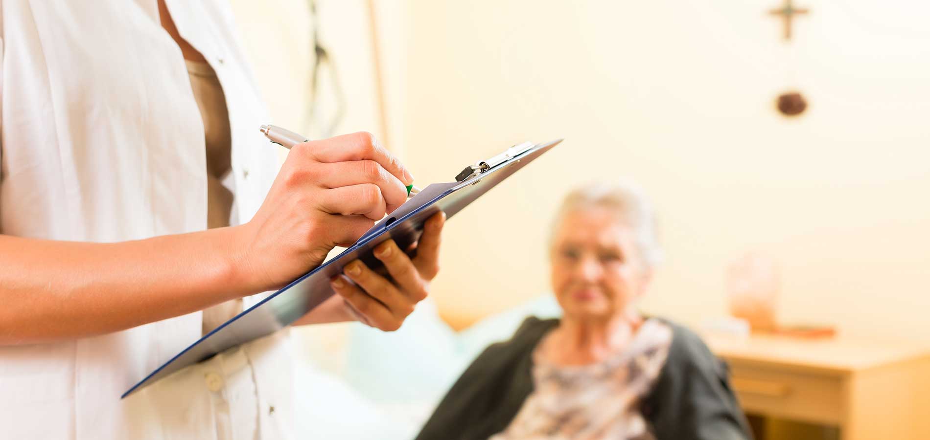 A healthcare worker writes on a clipboard with an elderly woman in the background sitting in a care home room.