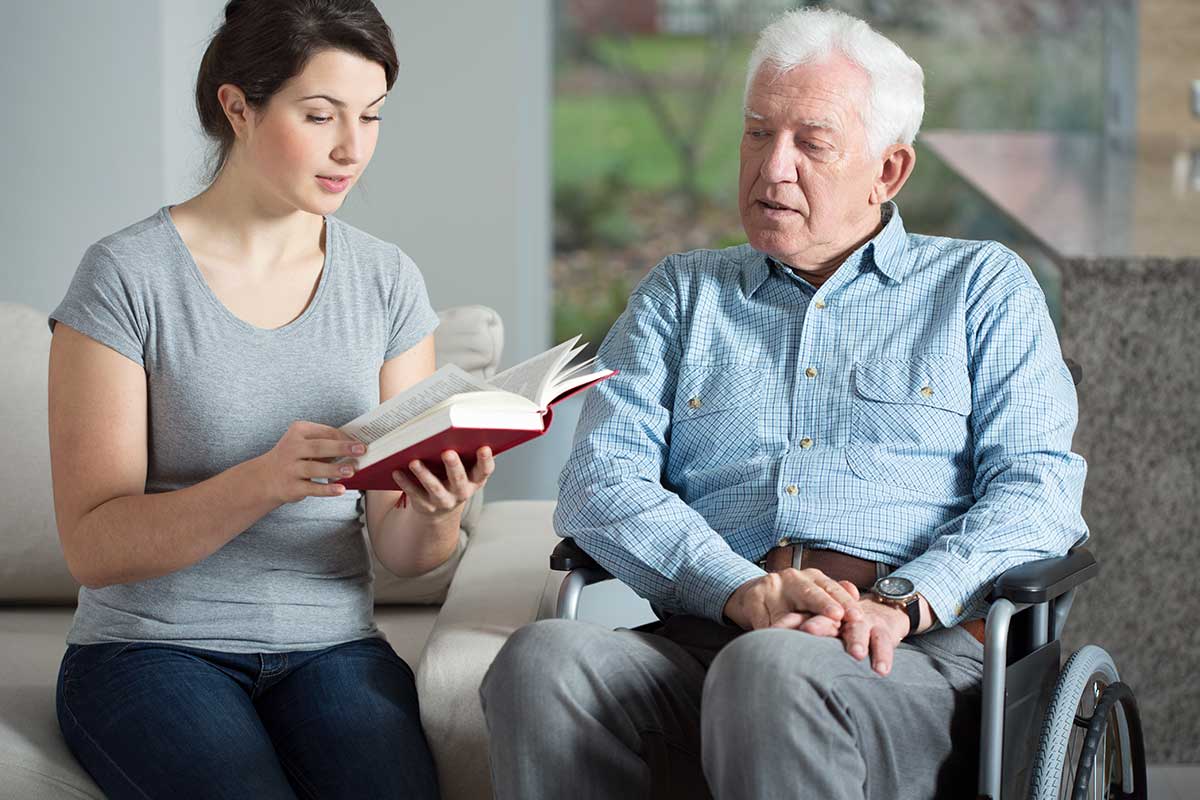 A young woman reads a book to an elderly man in a wheelchair in a living room.