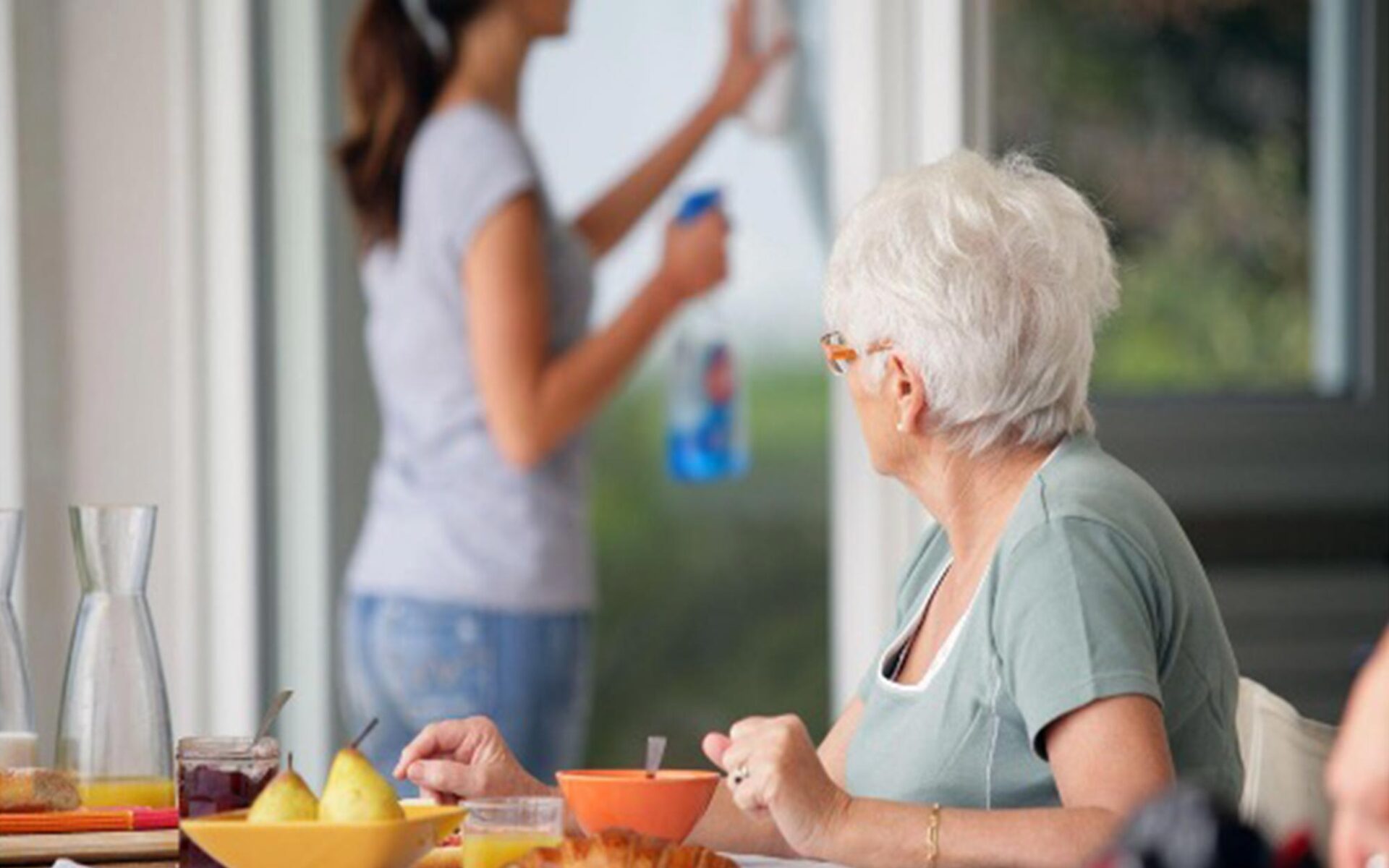 An elderly woman with white hair sits at a breakfast table, while a younger woman in the background cleans a window.