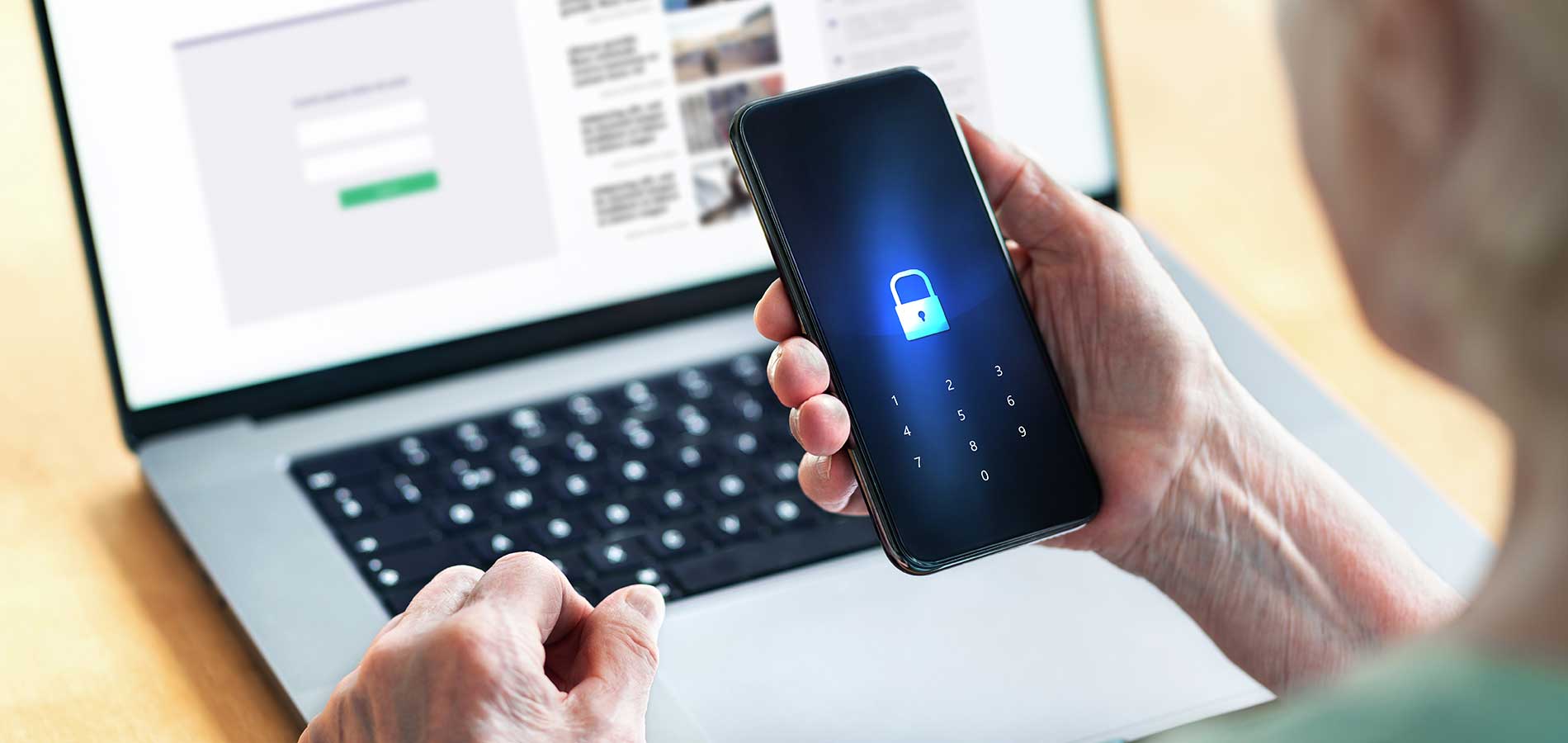 Person holding a smartphone displaying a lock icon, with a laptop showing login screens in the background.