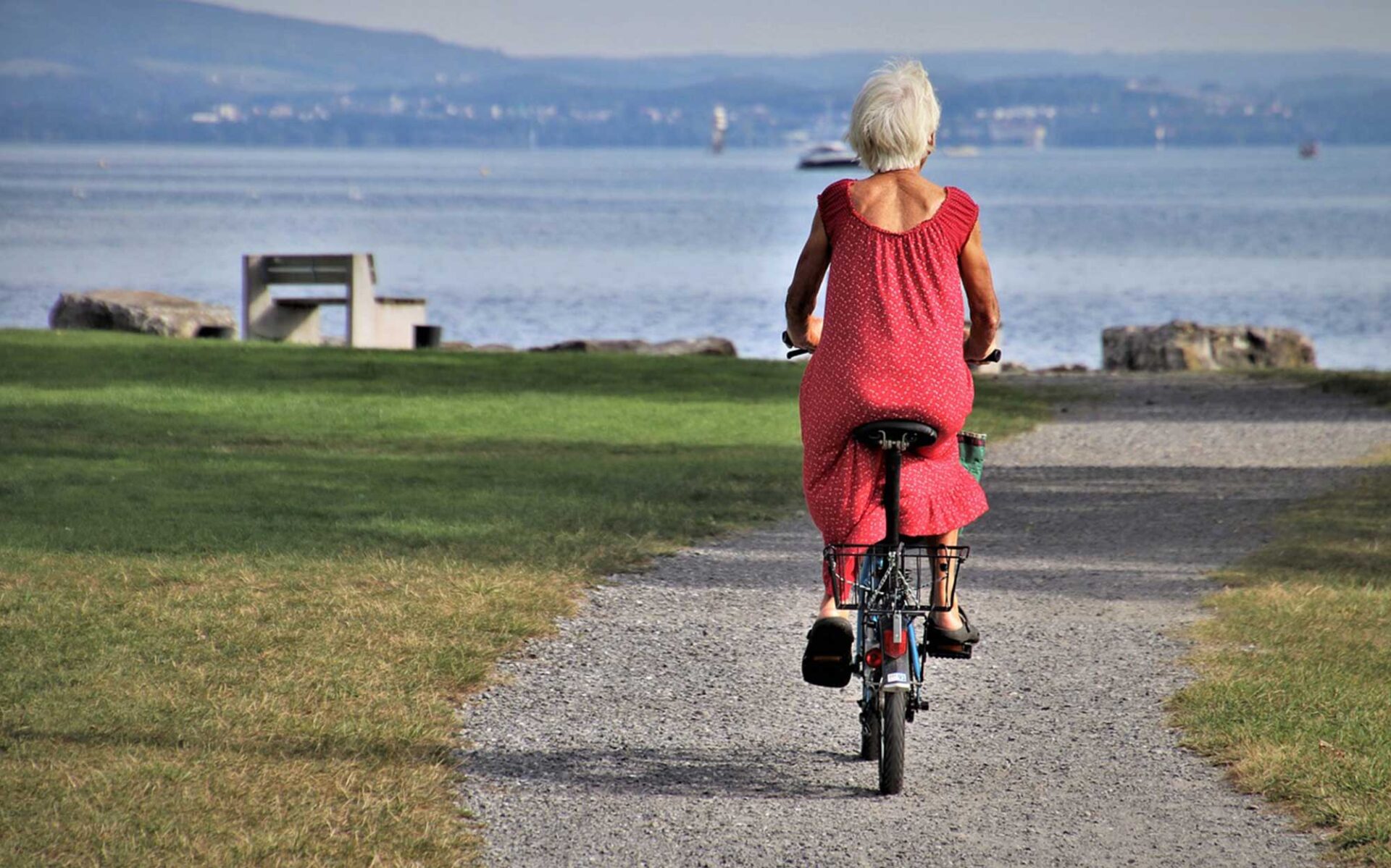 An elderly woman with white hair rides a bicycle down a gravel path towards a body of water, wearing a red dress.