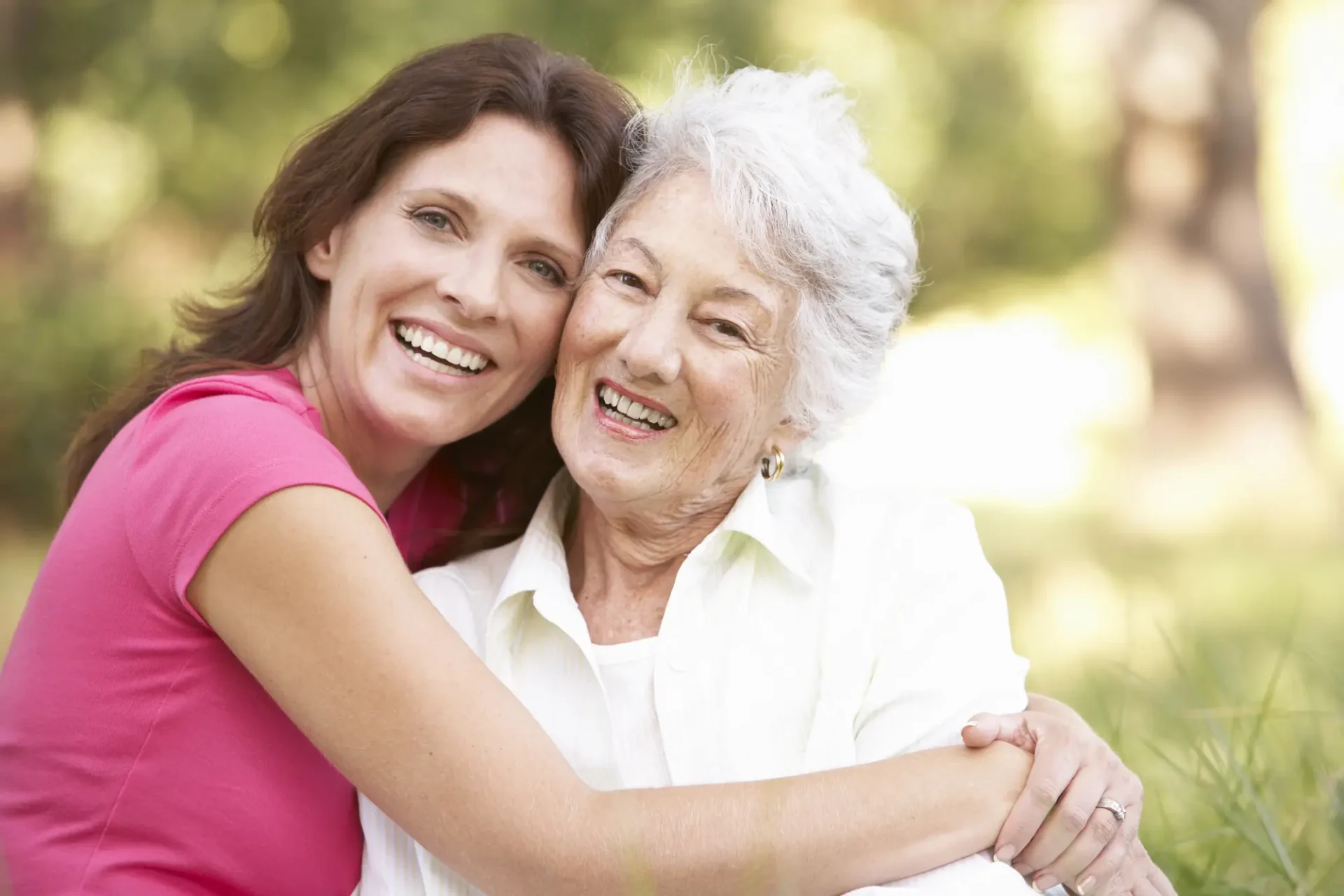 A younger woman and an older woman with gray hair smile and hug each other outdoors on a sunny day.