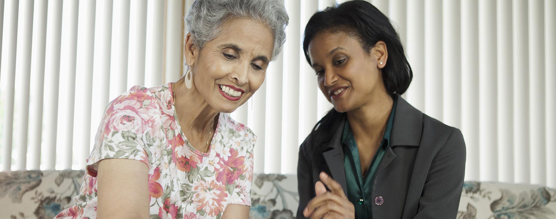 An elderly woman with gray hair and a floral blouse smiles while looking at a document that a younger woman in a dark blazer is pointing to, both sitting on a sofa.