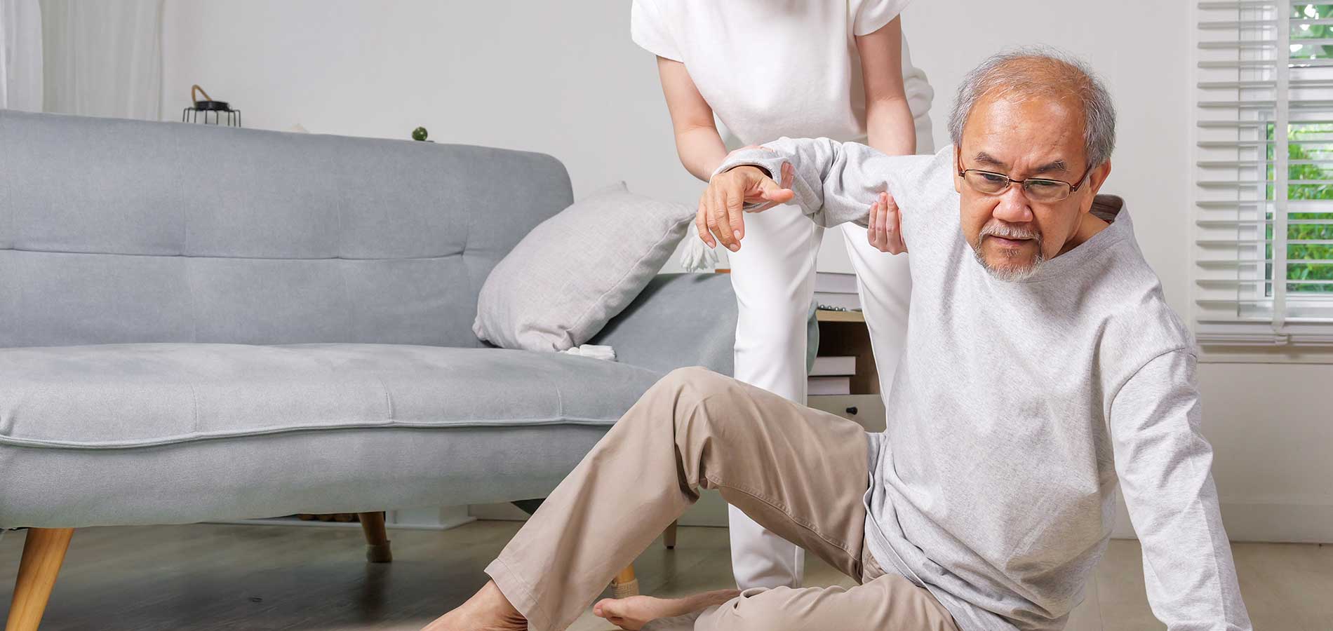 An elderly man being assisted by a woman as he tries to stand up from the floor next to a gray sofa in a bright living room.