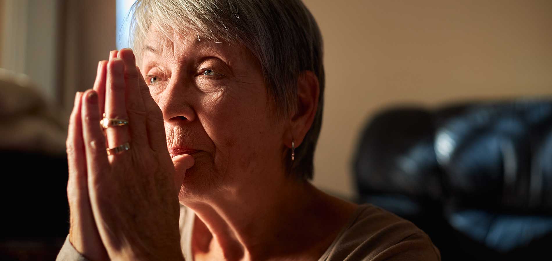 An elderly woman with short gray hair and rings on her fingers holds her hands together in a prayerful pose, gazing thoughtfully towards a light source.