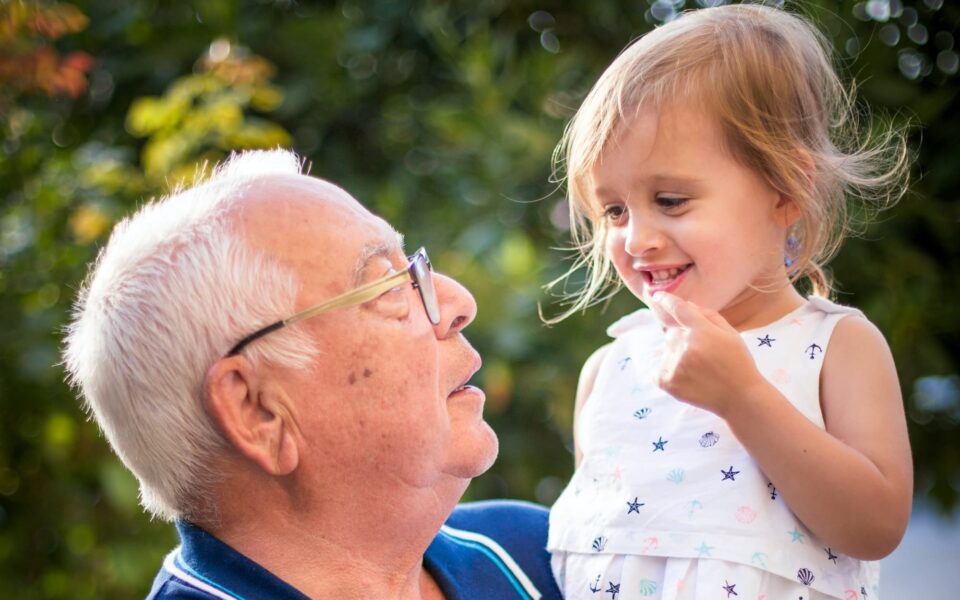 Grandfather holds young granddaughter as they smile at each other.