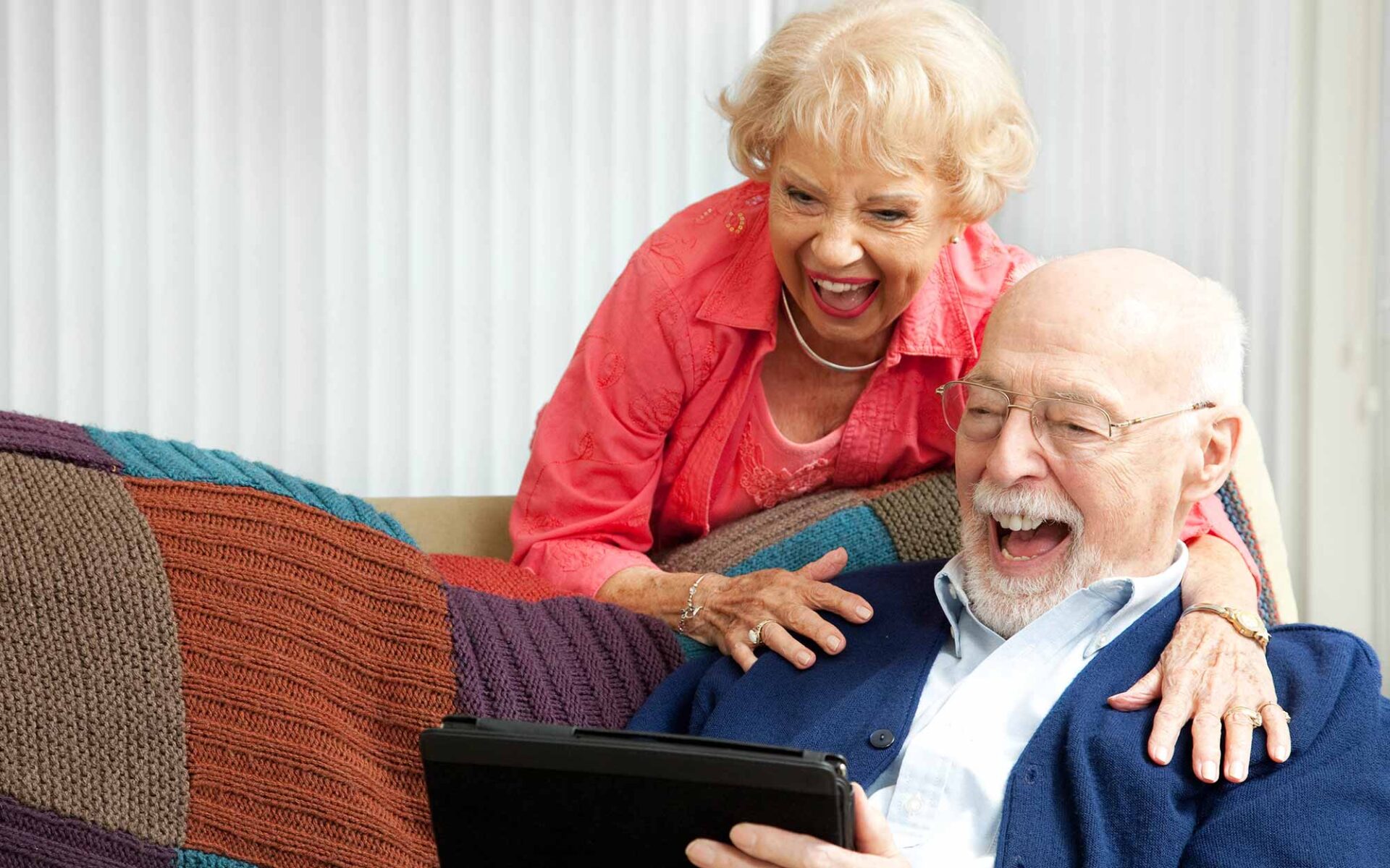 Elderly couple laughing together on a couch; the woman, in a pink blouse, stands behind the man, who is seated, holding a tablet.