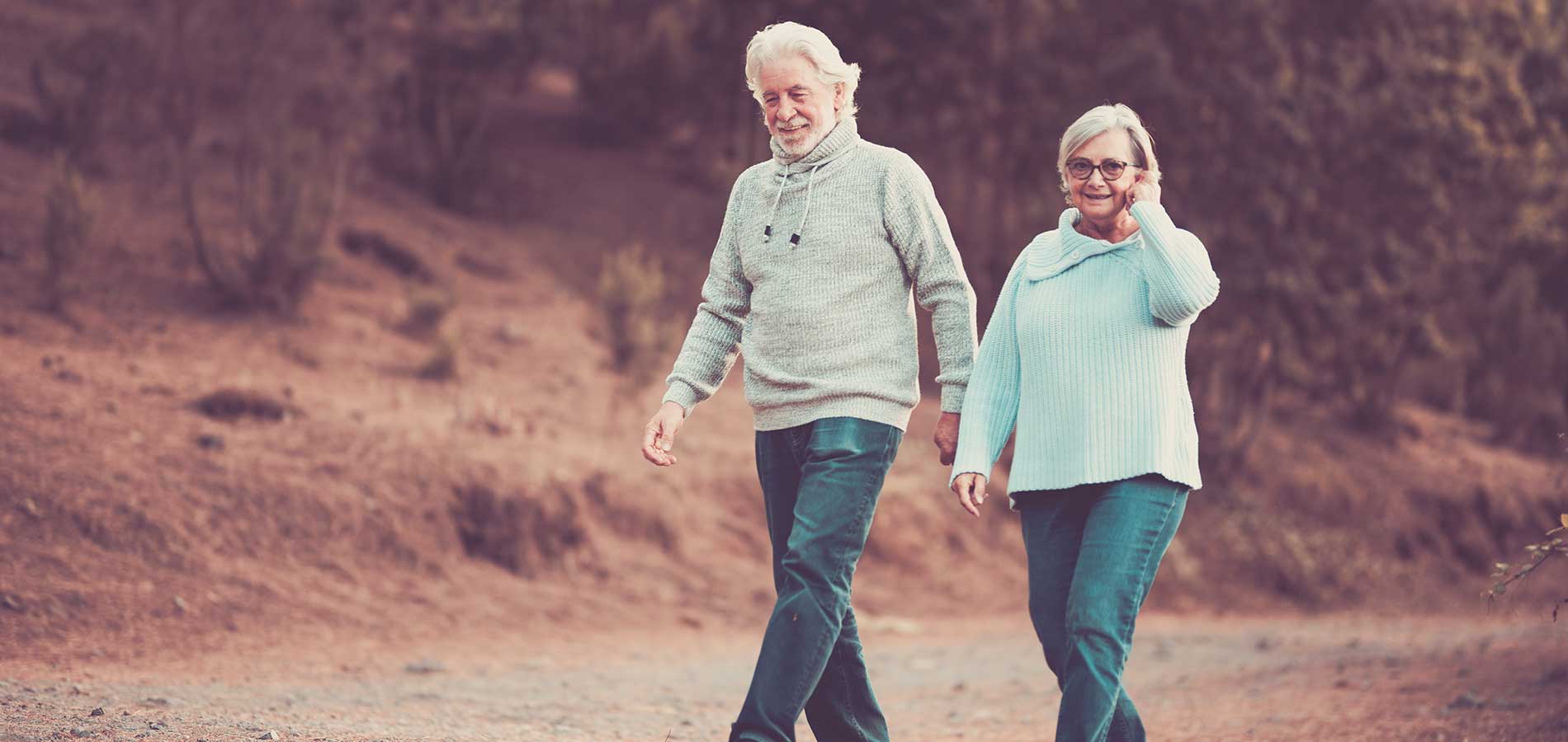 An elderly couple walking happily together on a dirt path through a natural setting.