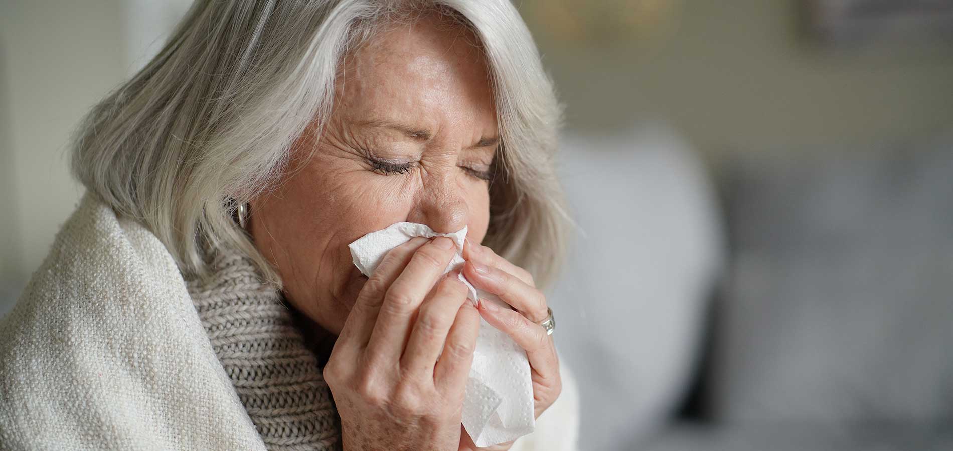 Elderly woman with white hair and a scarf, sneezing into a tissue, eyes closed, with a blurred background.