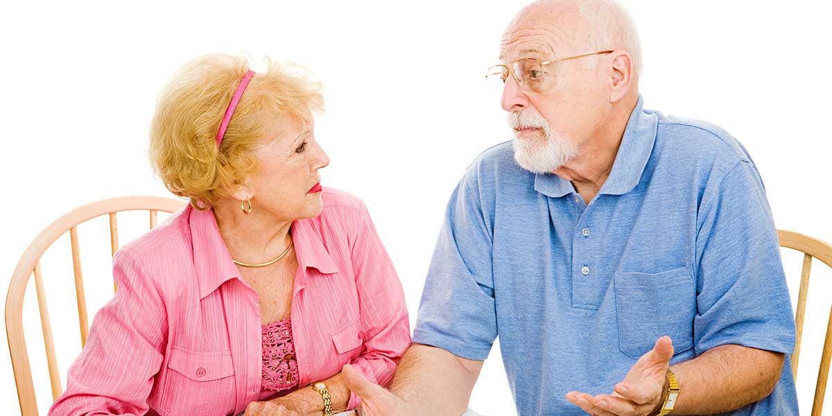 An elderly man in a blue shirt and a woman in a pink blouse conversing, sitting on wooden chairs against a white background.