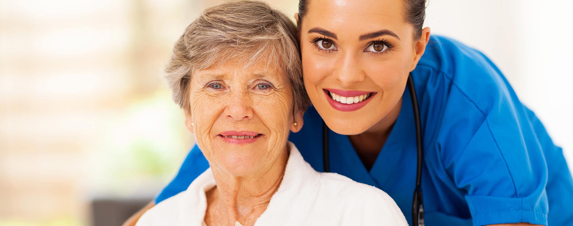 A smiling elderly woman and a cheerful nurse in blue scrubs posing closely for a photo.