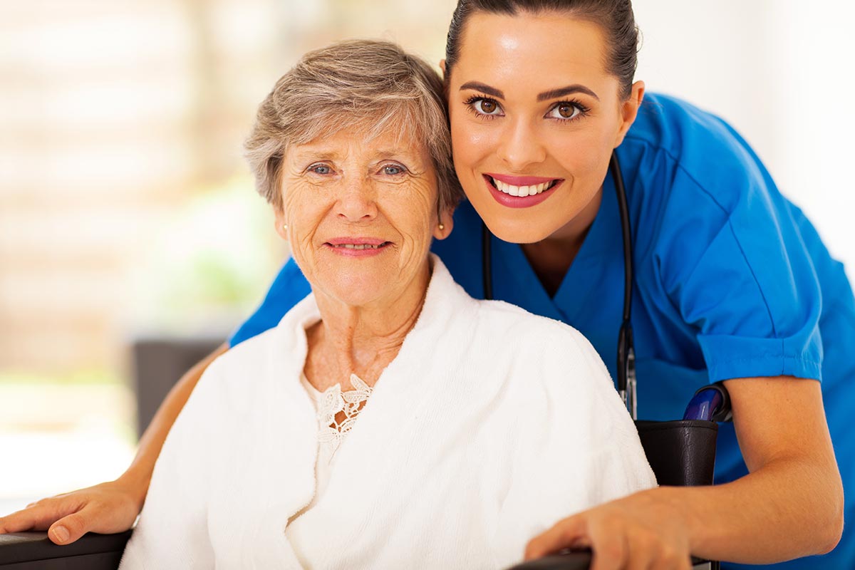 A smiling female nurse in blue scrubs posing with an elderly woman in a wheelchair indoors.