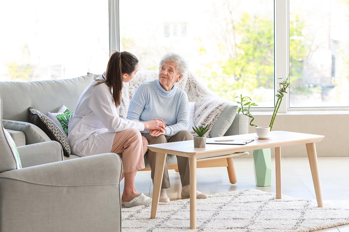 A nurse sitting on a sofa holds hands with an elderly woman in a bright living room, engaging in a conversation.