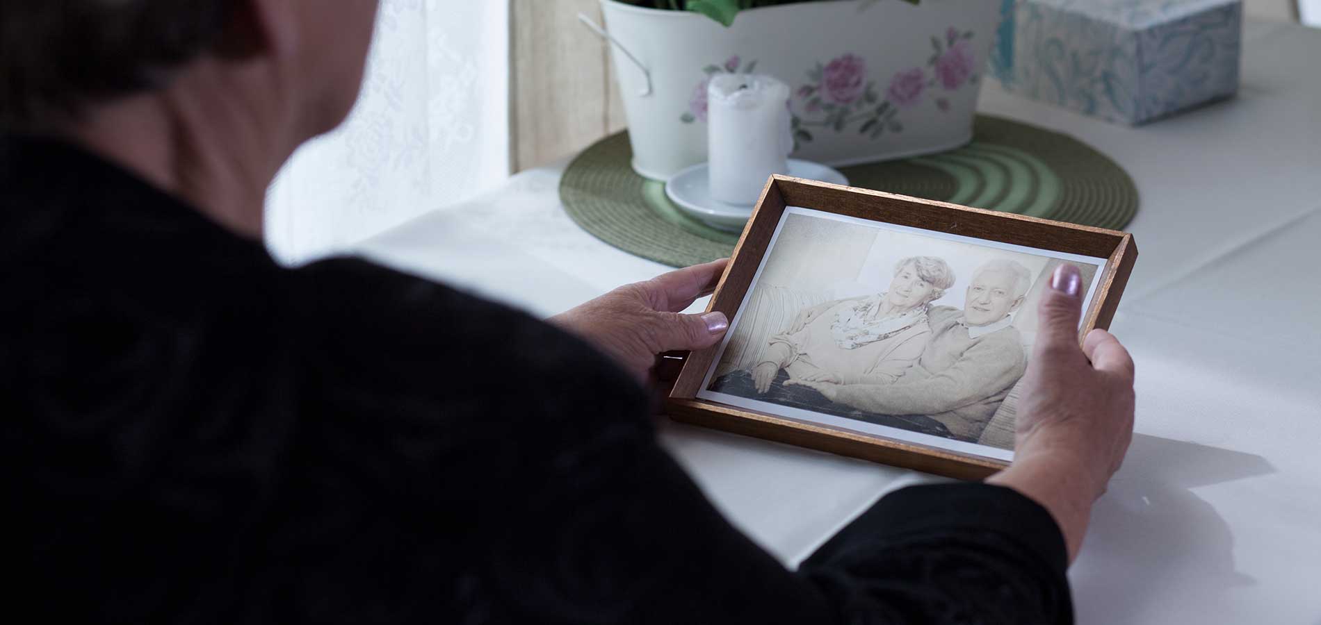 A woman looks at a framed photograph of an elderly couple, focusing on their smiling faces, at a dining table with a laid-back ambiance.