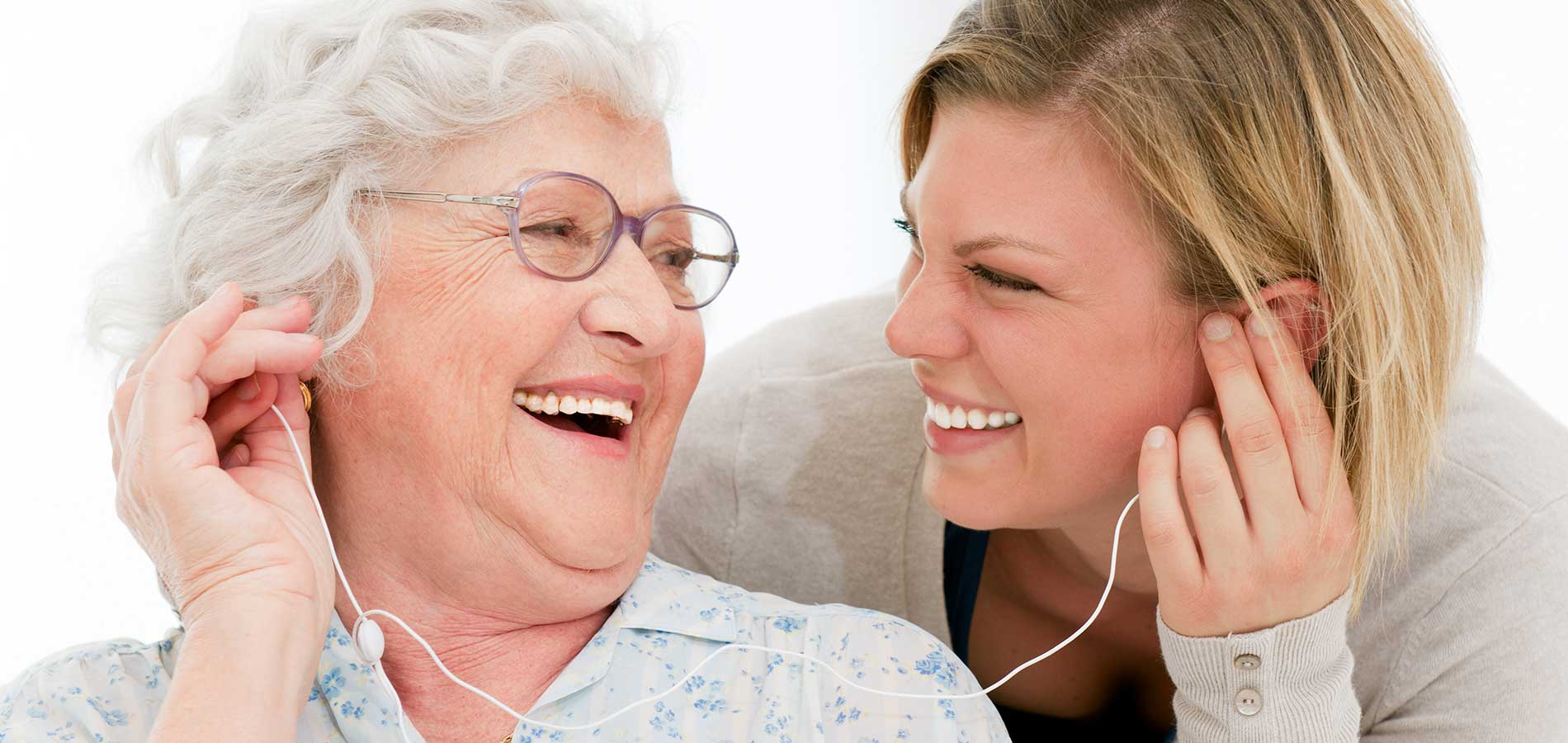 An elderly woman with glasses and a young woman smiling at each other while sharing earbuds, both wearing casual clothing.