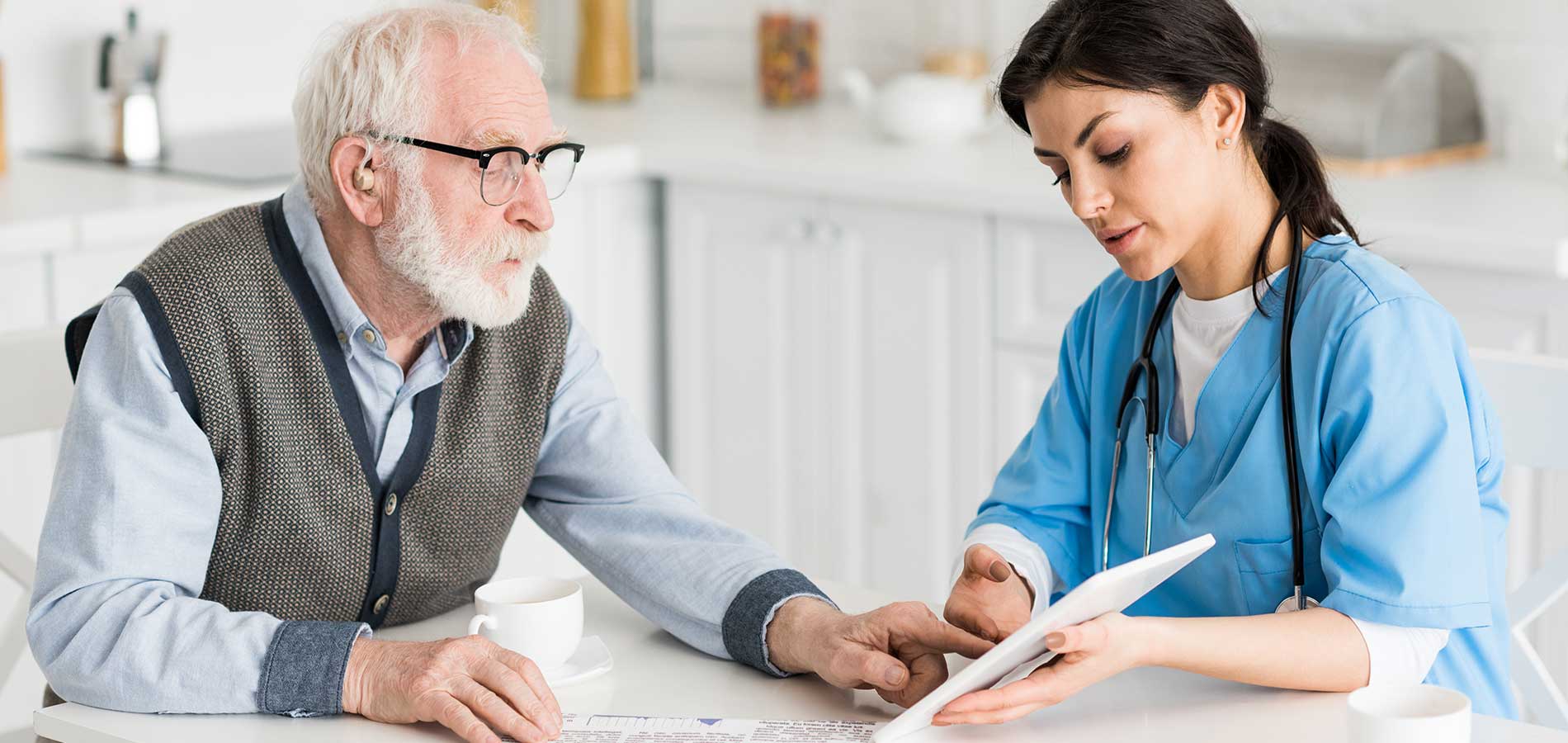 A nurse discusses medical documents with an elderly man at a kitchen table, both looking at a tablet.