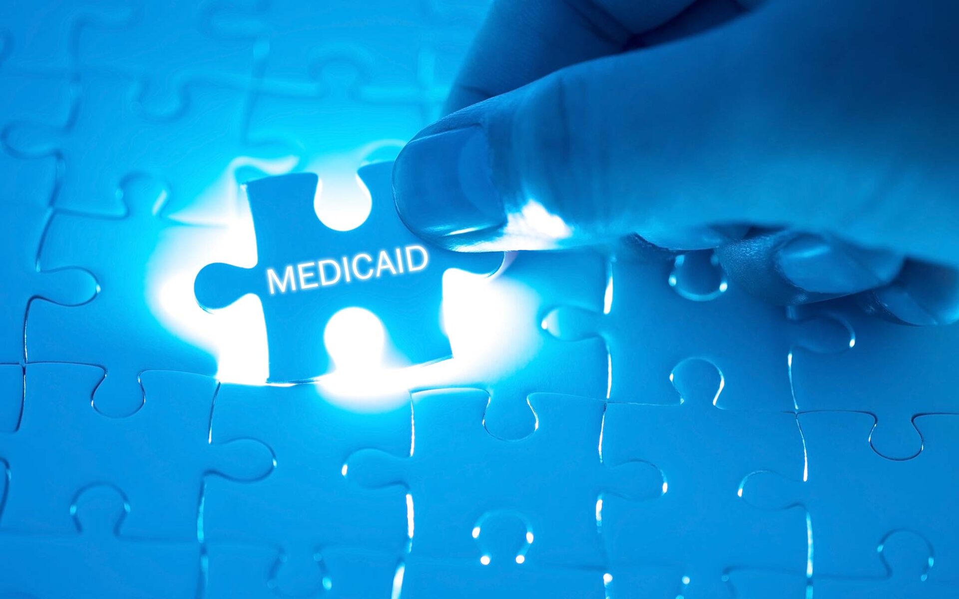 A hand places a puzzle piece labeled "Medicaid" into a puzzle, illuminated by blue light.