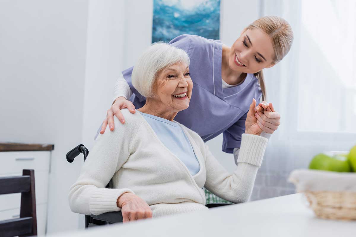 A caregiver standing beside an elderly woman in a wheelchair, both smiling joyfully in a bright, modern kitchen.