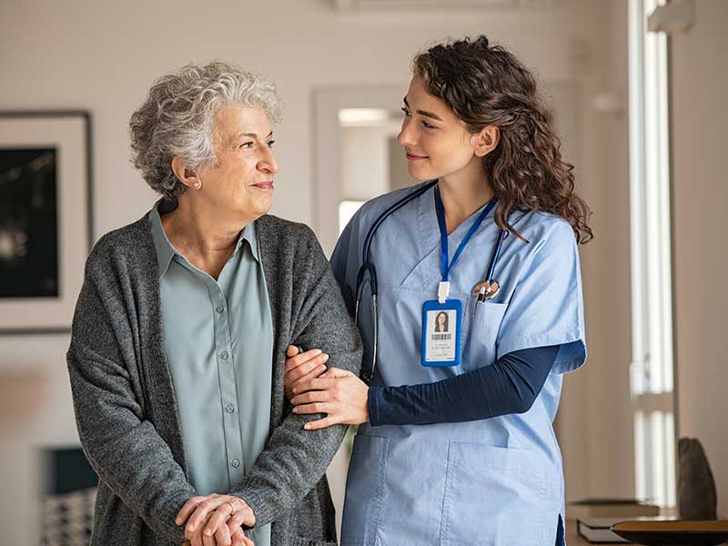 A nurse in blue scrubs assisting an elderly woman with curly gray hair as they walk through a hallway, both smiling warmly.