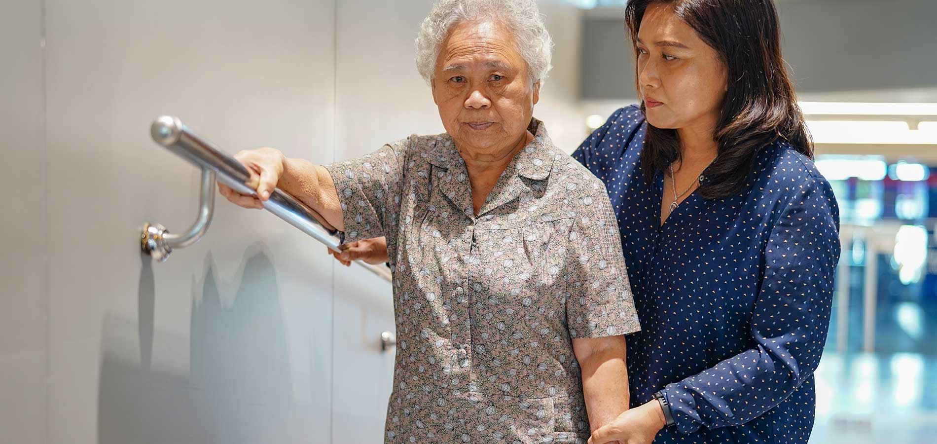 An elderly woman with a walking stick is assisted by a younger woman as they walk through a corridor.