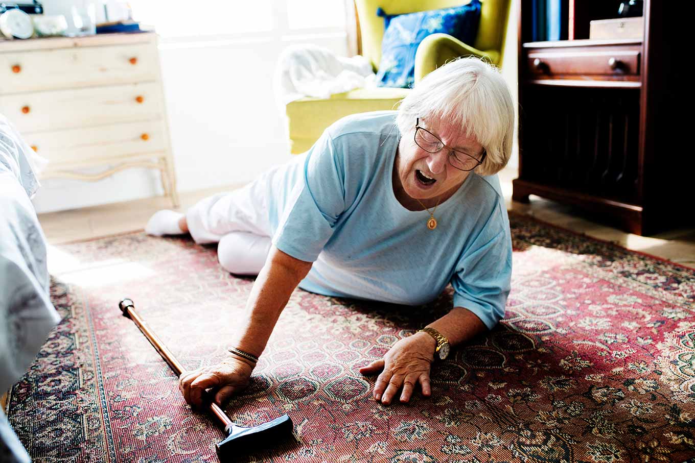 An elderly woman is lying on the floor, looking distressed, with a cane beside her on a patterned rug.