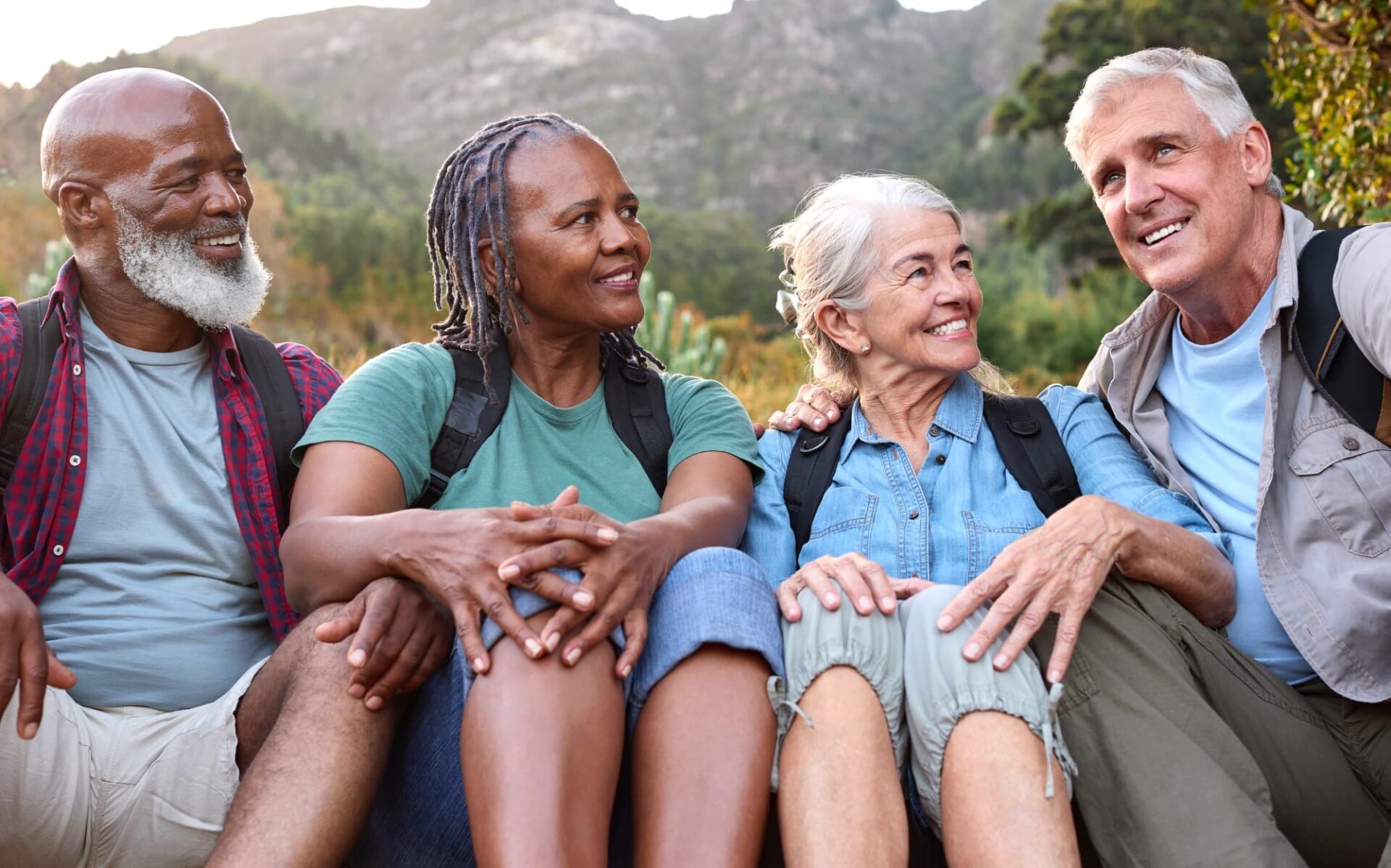 Four older adults, wearing casual outdoor clothing and backpacks, sit together outdoors with mountains in the background, smiling and talking to each other.