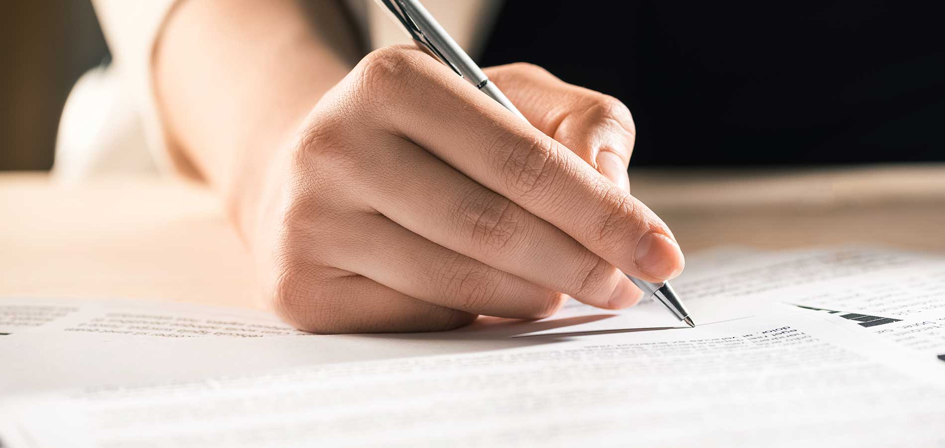 Person writing on a paper document with a pen, close-up on hand and pen over a well-lit desk.