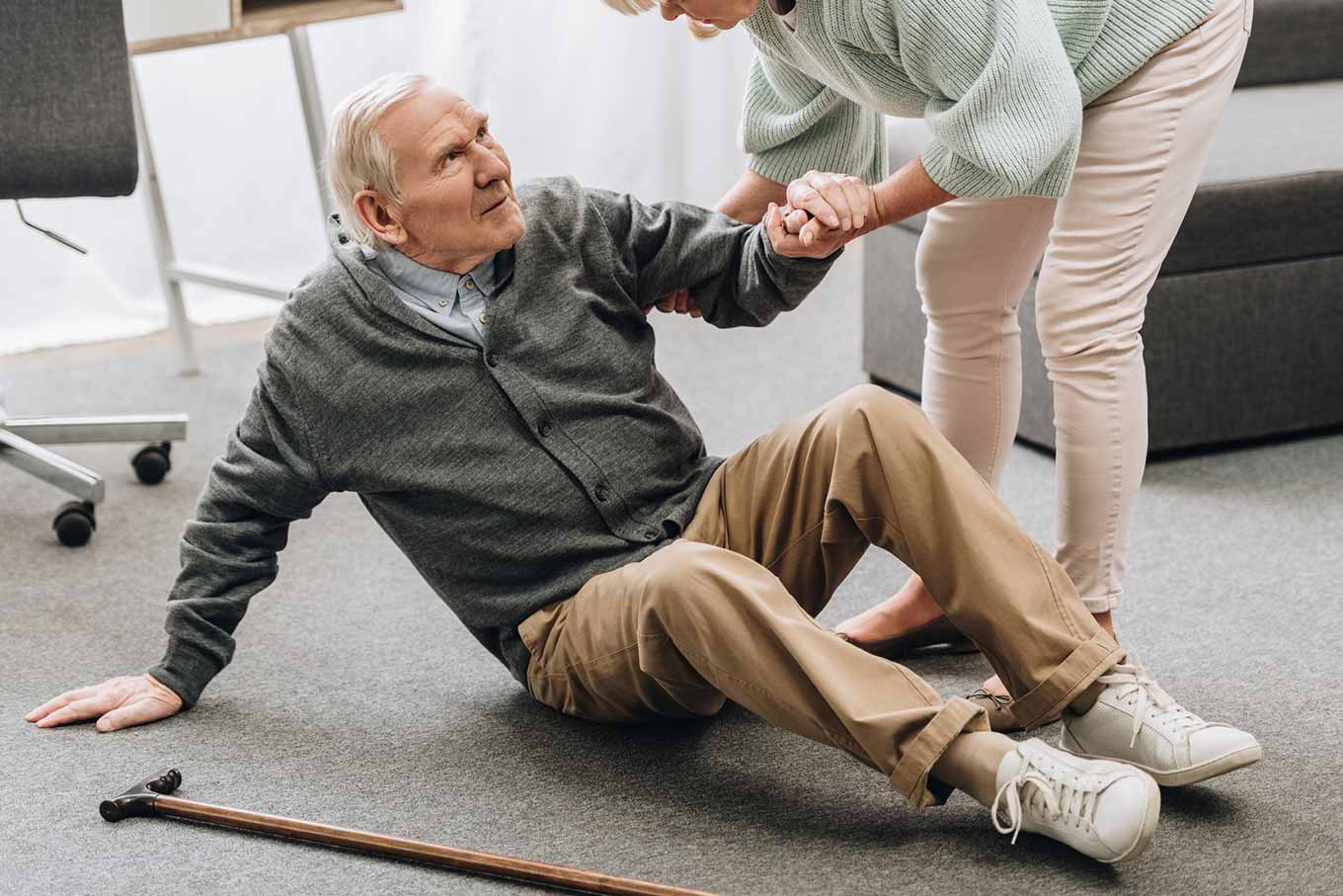 An elderly man sits on the floor holding his cane. A person assists him by the arm.