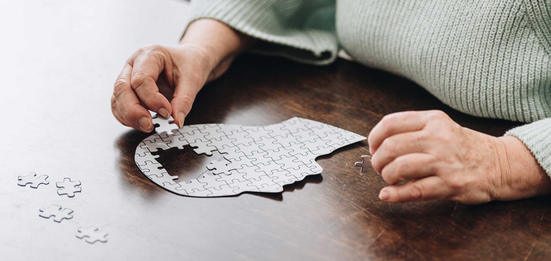 A person in a green sweater is assembling a jigsaw puzzle on a wooden table, focusing on placing a piece.