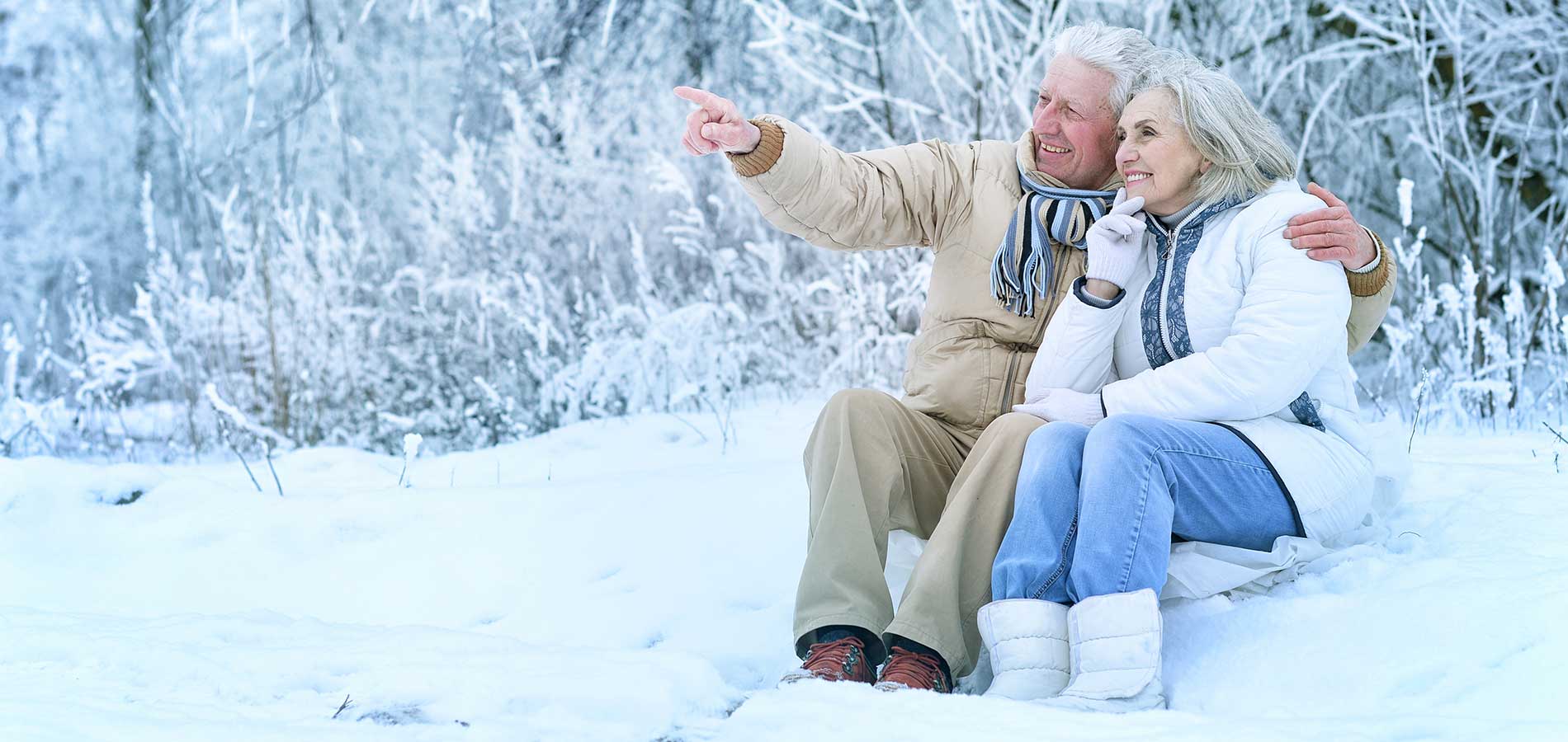 An elderly couple happily sitting in the snow, with the man pointing at something in the distance.