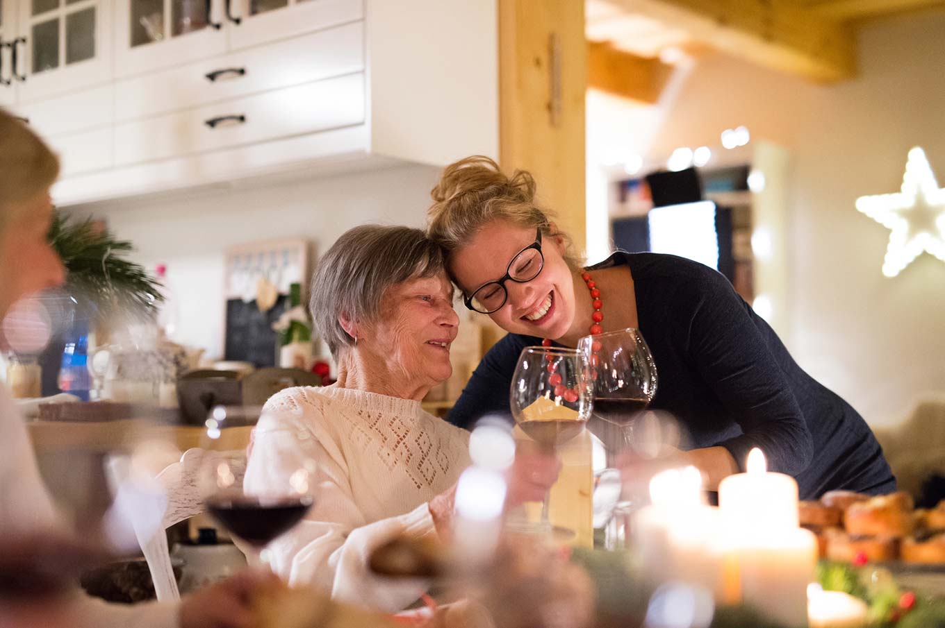 Two women share a joyful moment at a dinner table, surrounded by candles and wine glasses in a warmly lit room.