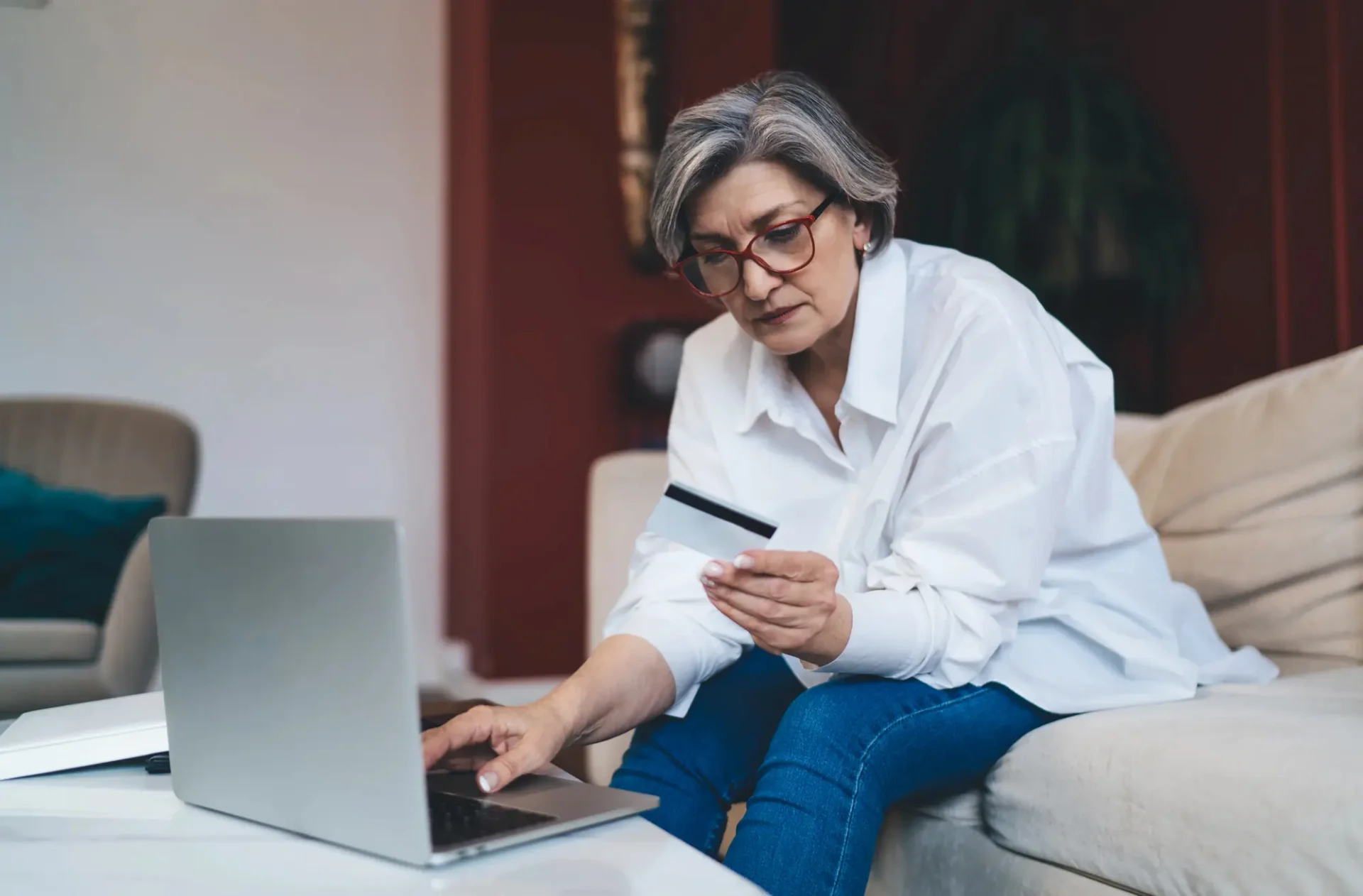 A woman sits on a couch, looking at a credit card in her hand while using a laptop on a table in front of her.